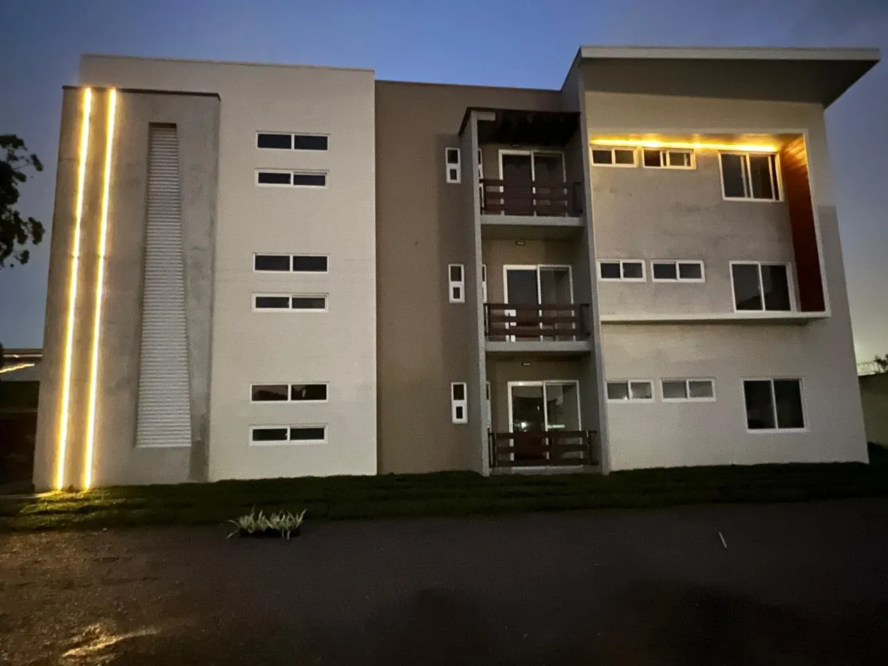Edificio de apartamentos de tres niveles al anochecer, con una franja de luz vertical en la fachada y los balcones iluminados.
